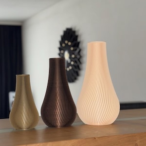 Può includere: Tre vasi decorativi di diverse dimensioni e colori. Il più piccolo è oro chiaro, quello centrale è marrone e il più grande è color pesca chiaro. Ogni vaso ha un design verticale a coste. Sono esposti su una superficie di legno.