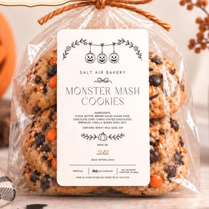 May include: A clear plastic bag tied with brown twine containing several round cookies with chocolate chips and orange candy-coated chocolates. A white rectangular label features the text Salt Air Bakery, Monster Mash Cookies, ingredients, and a date of 10/20.