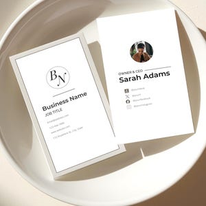 May include: Two white business cards with black text and a minimalist design are displayed in a white bowl. One card features a company logo and contact information. The other card displays the name "Sarah Adams" and social media icons.
