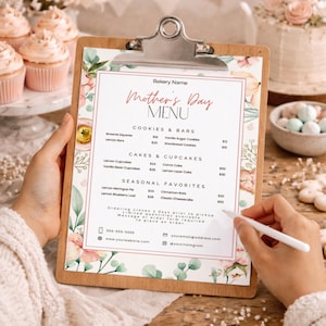 May include: A wooden clipboard with a floral Mother's Day menu. The menu lists cookies, cakes, and cupcakes. A hand holds a pen, ready to take an order. Cupcakes and a cake are in the background.