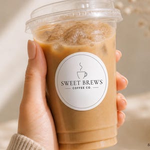 May include: A clear plastic cup filled with iced coffee, held by a person. The cup has a clear lid and a white circular sticker with the text "Sweet Brews Coffee Co." and a coffee cup graphic. The coffee is light brown with ice cubes.