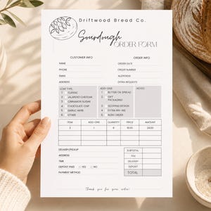 Puede incluir: Un formulario de pedido de masa madre blanco de Driftwood Bread Co. es sostenido por una persona. El formulario incluye secciones para información del cliente y del pedido, tipo de pan, complementos y notas. Una hogaza de pan y un cuenco de harina están en el fondo.