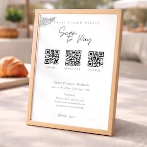 Puede incluir: Un letrero con marco de madera para Sweet Crumb Bakery con instrucciones de "Escanear para pagar". Muestra códigos QR para Venmo, Apple Pay y PayPal, junto con información de contacto y un mensaje de agradecimiento. El letrero está sobre una superficie clara.