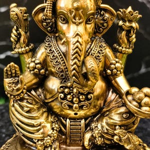 K&ouml;nnte beinhalten: Eine metallisch goldfarbene Statue der Gottheit Ganesha, dargestellt in sitzender Position mit vier Armen. Die Figur zeigt einen Elefantenkopf, verzierten Schmuck und eine Krone. Eine Hand zeigt ein Om-Symbol, w&auml;hrend eine andere eine Schale mit runden Objekten h&auml;lt.