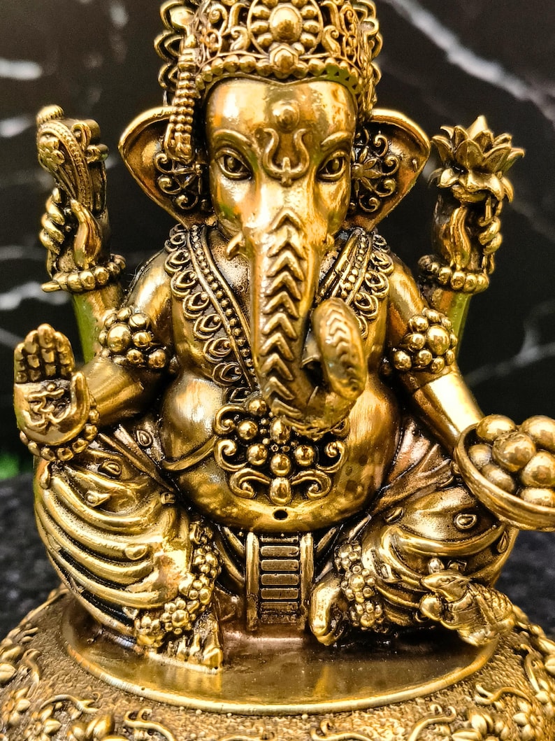 K&ouml;nnte beinhalten: Eine metallisch goldfarbene Statue der Gottheit Ganesha, dargestellt in sitzender Position mit vier Armen. Die Figur zeigt einen Elefantenkopf, verzierten Schmuck und eine Krone. Eine Hand zeigt ein Om-Symbol, w&auml;hrend eine andere eine Schale mit runden Objekten h&auml;lt.