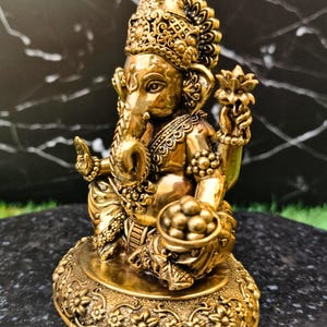 K&ouml;nnte beinhalten: Eine goldfarbene Metallstatue der Gottheit Ganesha in sitzender Position. Die Figur zeigt einen Elefantenkopf, verzierten Schmuck und eine Krone. Eine Hand ist zum Segen erhoben, w&auml;hrend die andere eine Schale mit runden S&uuml;&szlig;igkeiten h&auml;lt.