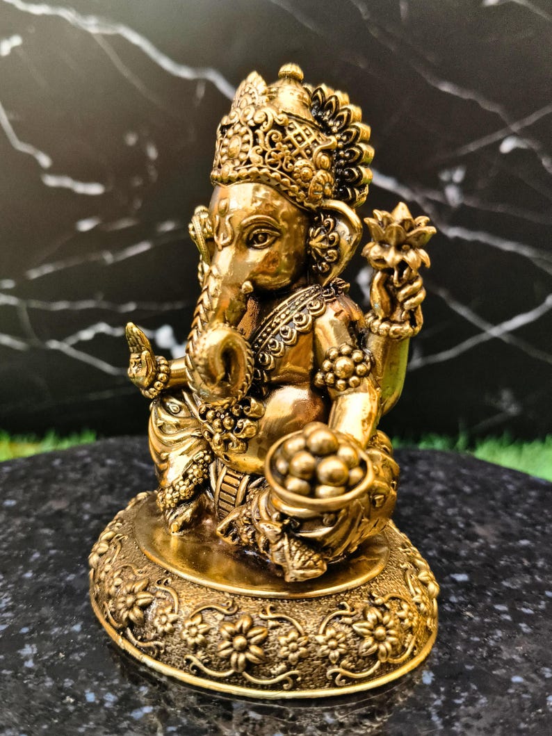 K&ouml;nnte beinhalten: Eine goldfarbene Metallstatue der Gottheit Ganesha in sitzender Position. Die Figur zeigt einen Elefantenkopf, verzierten Schmuck und eine Krone. Eine Hand ist zum Segen erhoben, w&auml;hrend die andere eine Schale mit runden S&uuml;&szlig;igkeiten h&auml;lt.