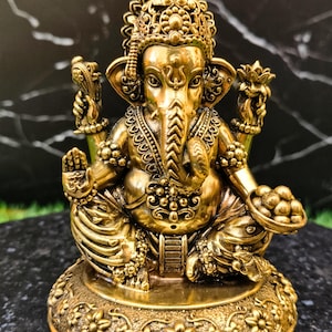 K&ouml;nnte beinhalten: Eine goldfarbene Metallstatue der Gottheit Ganesha, sitzend mit vier Armen dargestellt. Die Figur hat einen Elefantenkopf, verzierten Schmuck, eine Krone und h&auml;lt eine Schale mit runden S&uuml;&szlig;igkeiten in einer Hand, auf einem Blumenfu&szlig;.
