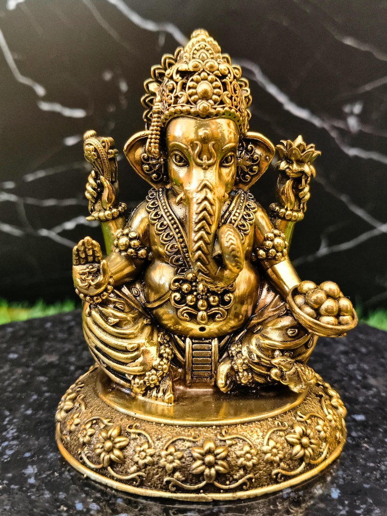 K&ouml;nnte beinhalten: Eine goldfarbene Metallstatue der Gottheit Ganesha, sitzend mit vier Armen dargestellt. Die Figur hat einen Elefantenkopf, verzierten Schmuck, eine Krone und h&auml;lt eine Schale mit runden S&uuml;&szlig;igkeiten in einer Hand, auf einem Blumenfu&szlig;.