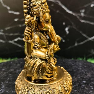 K&ouml;nnte beinhalten: Eine metallisch goldfarbene Figur der Gottheit Ganesha in sitzender Position. Die Figur zeigt einen Elefantenkopf, mehrere Arme und detaillierten Schmuck und ruht auf einem runden Sockel, der mit einem floralen Reliefmuster verziert ist.