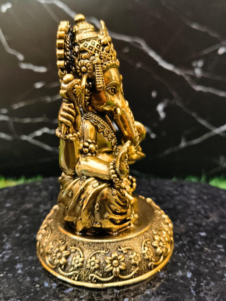K&ouml;nnte beinhalten: Eine metallisch goldfarbene Figur der Gottheit Ganesha in sitzender Position. Die Figur zeigt einen Elefantenkopf, mehrere Arme und detaillierten Schmuck und ruht auf einem runden Sockel, der mit einem floralen Reliefmuster verziert ist.