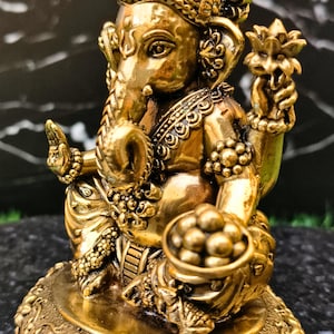 K&ouml;nnte beinhalten: Eine goldfarbene Metallstatue der Gottheit Ganesha mit Elefantenkopf und vier Armen. Die Figur tr&auml;gt eine verzierte Krone sowie Schmuck, h&auml;lt eine Lotusbl&uuml;te und eine Schale mit runden S&uuml;&szlig;igkeiten und sitzt auf einem runden Blumensockel.