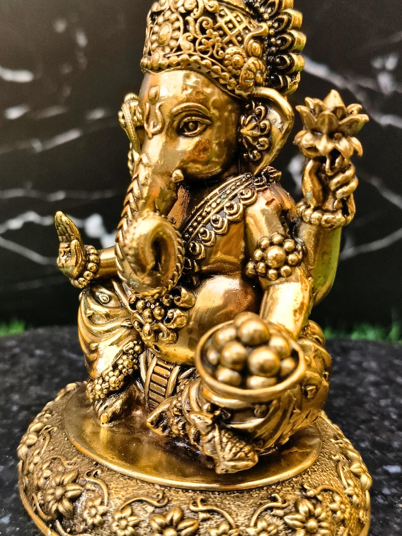 K&ouml;nnte beinhalten: Eine goldfarbene Metallstatue der Gottheit Ganesha mit Elefantenkopf und vier Armen. Die Figur tr&auml;gt eine verzierte Krone sowie Schmuck, h&auml;lt eine Lotusbl&uuml;te und eine Schale mit runden S&uuml;&szlig;igkeiten und sitzt auf einem runden Blumensockel.