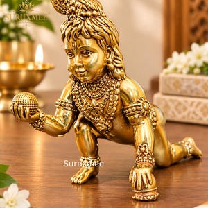 May include: A polished brass statue of a crawling child deity, featuring intricate jewellery, a textured ball held in the right hand, and a peacock feather ornament in the hair. The figure is depicted in a kneeling posture with the text Suruxmee visible.