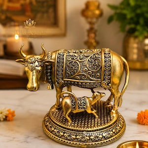 May include: A gold-toned metallic figurine depicting a cow and a calf standing on a circular, textured base. The cow features intricate embossed floral patterns on its back covering and head, while the calf has a matching decorative blanket.