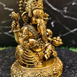 K&ouml;nnte beinhalten: Eine goldfarbene Metallstatue der Gottheit Ganesha mit Elefantenkopf und vier Armen. Die Figur sitzt im Schneidersitz auf einem runden Sockel mit floralen Verzierungen und tr&auml;gt kunstvollen Schmuck sowie eine Krone auf dem Kopf.