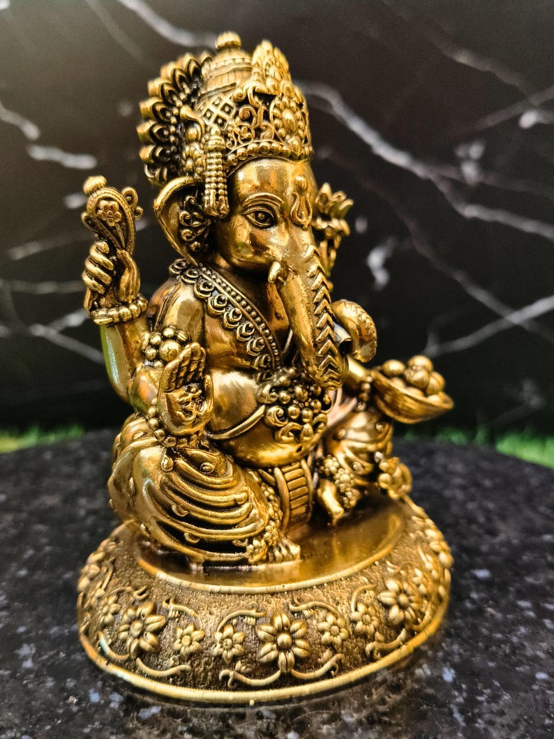 K&ouml;nnte beinhalten: Eine goldfarbene Metallstatue der Gottheit Ganesha mit Elefantenkopf und vier Armen. Die Figur sitzt im Schneidersitz auf einem runden Sockel mit floralen Verzierungen und tr&auml;gt kunstvollen Schmuck sowie eine Krone auf dem Kopf.