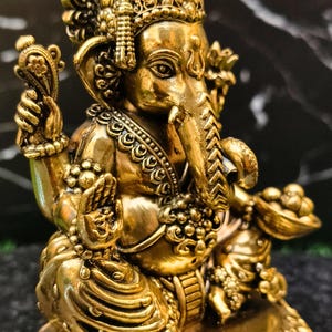 K&ouml;nnte beinhalten: Eine goldfarbene Metallstatue der Gottheit Ganesha in sitzender Position. Die Figur zeigt einen Elefantenkopf, mehrere Arme und aufwendige ornamentale Details, darunter eine Krone, Halsketten und eine Schale, die in einer Hand gehalten wird.