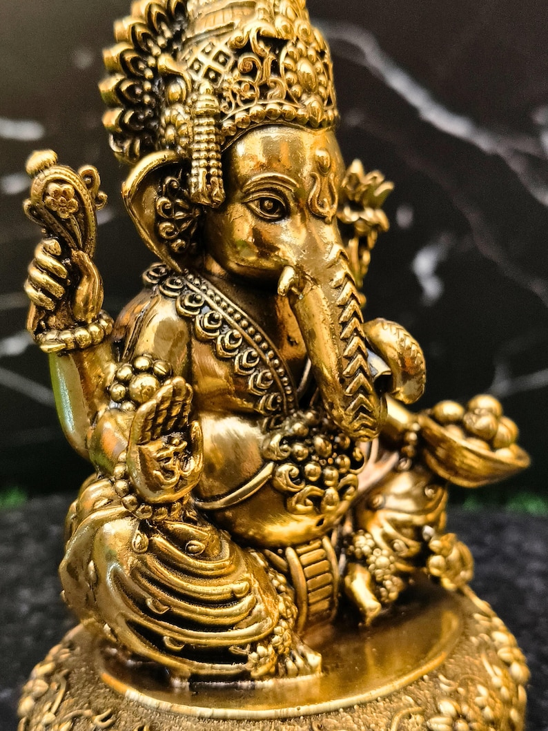 K&ouml;nnte beinhalten: Eine goldfarbene Metallstatue der Gottheit Ganesha in sitzender Position. Die Figur zeigt einen Elefantenkopf, mehrere Arme und aufwendige ornamentale Details, darunter eine Krone, Halsketten und eine Schale, die in einer Hand gehalten wird.