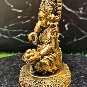 K&ouml;nnte beinhalten: Eine goldfarbene Metallstatue der Gottheit Ganesha in sitzender Position mit gekreuzten Beinen. Die Figur hat einen Elefantenkopf, eine Krone und h&auml;lt eine Lotusbl&uuml;te in der einen und eine Schale mit runden Opfergaben in der anderen Hand.