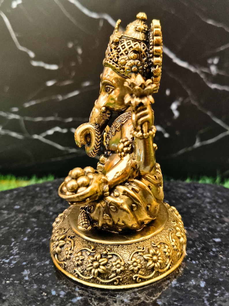 K&ouml;nnte beinhalten: Eine goldfarbene Metallstatue der Gottheit Ganesha in sitzender Position mit gekreuzten Beinen. Die Figur hat einen Elefantenkopf, eine Krone und h&auml;lt eine Lotusbl&uuml;te in der einen und eine Schale mit runden Opfergaben in der anderen Hand.