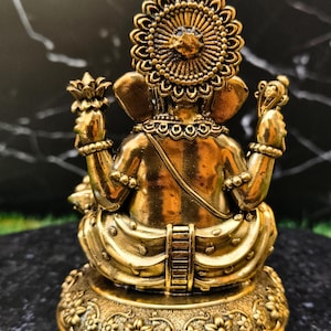 K&ouml;nnte beinhalten: Eine metallisch goldfarbene Figur einer Gottheit von hinten gesehen, mit einem gro&szlig;en kreisf&ouml;rmigen Heiligenschein, vier Armen mit erhobenen H&auml;nden, die Gegenst&auml;nde halten, und einer sitzenden Haltung auf einem runden Sockel mit floralen Schnitzereien.