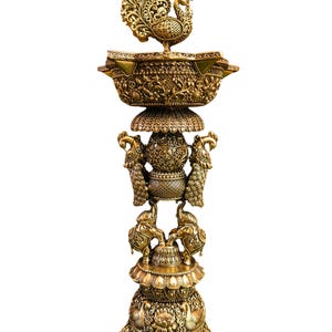 May include: A tall, ornate brass oil lamp featuring a peacock finial at the top, a central pillar adorned with two flanking peacocks, and a base supported by two elephants. The entire structure is covered in intricate, embossed floral patterns.