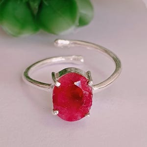 May include: A silver ring with a striking, oval-shaped, deep red gemstone. The open band design allows for size adjustment. The gemstone is held in place by four prongs. The ring is displayed on a soft, light surface. The ring's design is simple yet elegant.