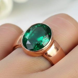 May include: A rose gold ring featuring a large, oval-cut emerald gemstone. The emerald is a deep, vibrant green, set in a polished, smooth band. The ring is shown on a finger, highlighting the gemstone's brilliance.