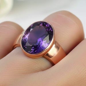 May include: A rose gold ring with a large, oval amethyst gemstone. The deep purple stone is set in a simple, polished band. The ring is shown on a finger, highlighting the gemstone's colour and clarity.