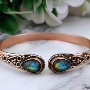Puede incluir: Pulsera de puño abierta con acabado cobrizo. Los extremos presentan detalles ornamentados y piedras en forma de lágrima con iridiscencia azul y amarilla. La pulsera se exhibe sobre una superficie de mármol.