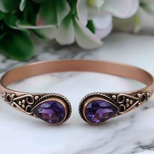 Op de afbeelding: Open manchetarmband met een koperen afwerking. De armband heeft twee druppelvormige, dieppaarse edelstenen. De uiteinden van de armband hebben sierlijke, gedetailleerde ontwerpen. De achtergrond is wazig, met witte en groene bladeren.