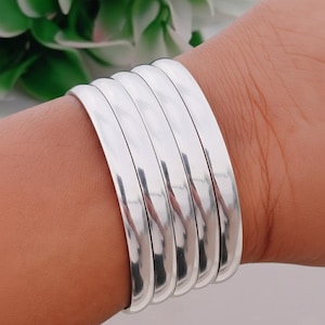 Puede incluir: Un conjunto de cinco pulseras plateadas pulidas. Las pulseras tienen un diseño liso y plano con bordes redondeados. Están apiladas en una muñeca, reflejando la luz. Cada pulsera mide aproximadamente 0,5 cm de ancho. Un accesorio sencillo y elegante.