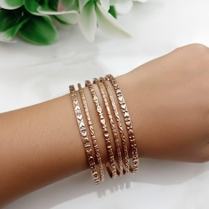 Può includere: Una collezione di sette braccialetti color rame, ognuno con un design inciso unico. I motivi includono cuori, motivi floreali e forme geometriche. I braccialetti sono impilati su un polso, mostrando le loro diverse trame e stili.