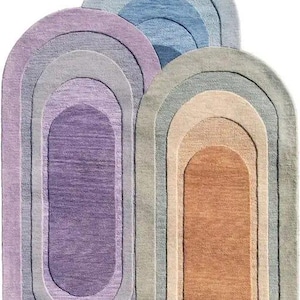 Puede incluir: Tres alfombras ovaladas en tonos morado, gris, azul y beige. Cada alfombra presenta un diseño en capas con diferentes tonos de color, creando un efecto degradado. Las alfombras están hechas de un material suave y texturizado.