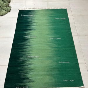 May include: A rectangular green rug with a gradient design, transitioning from a dark to a lighter shade of green. The rug has a fringe on the shorter ends and the text "Immo carpet" is printed on the rug.