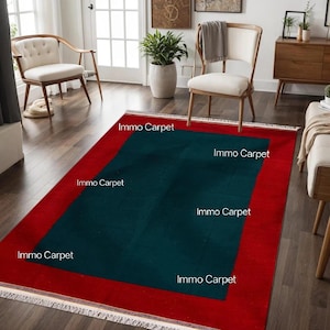 May include: A rectangular rug with a red border and a dark teal center. The rug is placed on a hardwood floor in a room with chairs and a plant. The text "Immo Carpet" is printed on the rug.