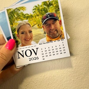 May include: A small calendar for November 2026, featuring a photograph of two people smiling. The calendar is held by a person with pink nail polish. The background shows a tropical scene with palm trees.