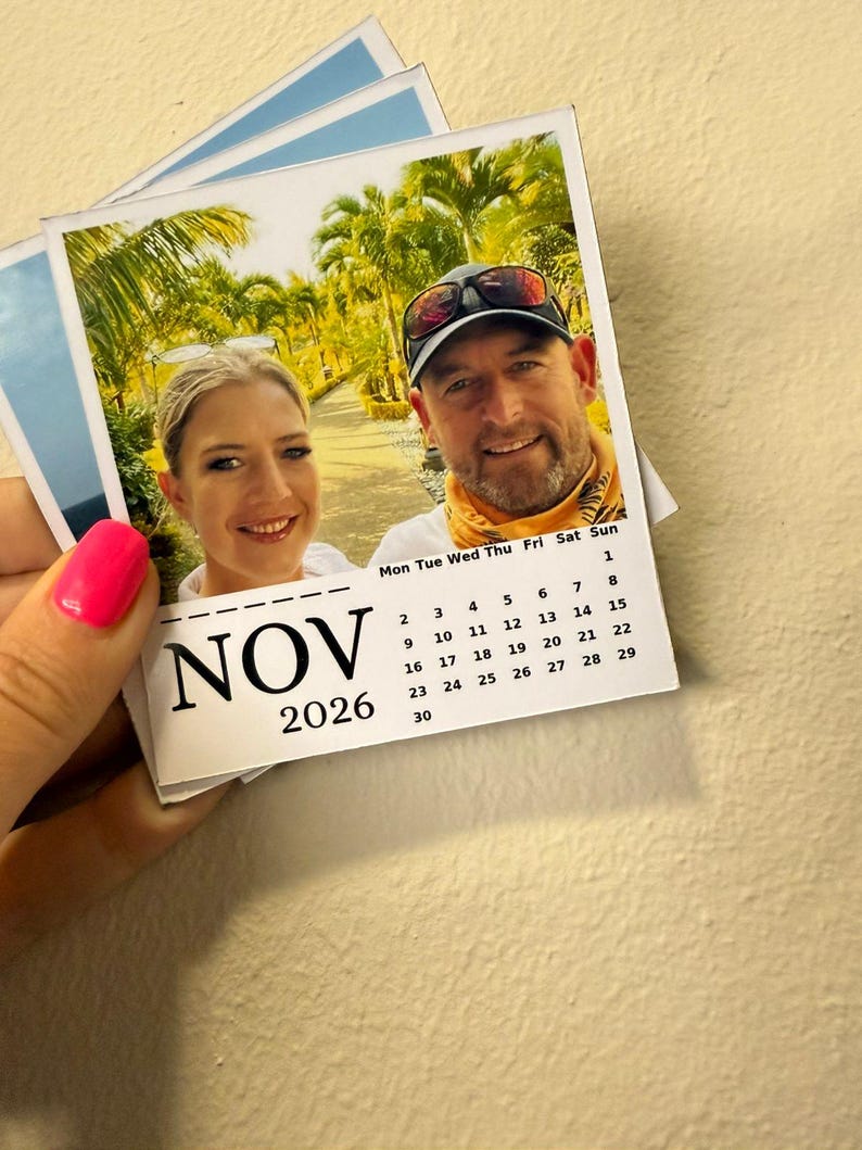 May include: A small calendar for November 2026, featuring a photograph of two people smiling. The calendar is held by a person with pink nail polish. The background shows a tropical scene with palm trees.