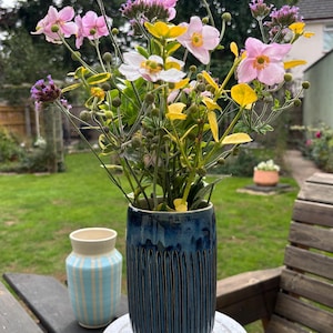 May include: A cylindrical dark blue ceramic vase with vertical ribbed texture containing a bouquet of pink anemone flowers, yellow buttercups, and purple verbena. A smaller light blue and white striped ceramic vase sits in the background.