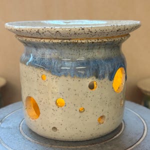 Puede incluir: Un quemador de aceite de cerámica moteada con un esmalte azul claro y crema. El calentador en forma de jarra tiene varias aberturas redondas y un borde ancho. Las aberturas brillan con una luz cálida.