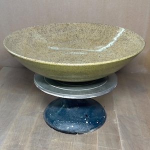 Puede incluir: Un cuenco grande de cerámica hecho a mano con un esmalte moteado marrón y marrón. El cuenco tiene una forma ancha y poco profunda y se asienta sobre un pedestal azul oscuro con motas blancas. El cuenco se exhibe en un torno de alfarero.