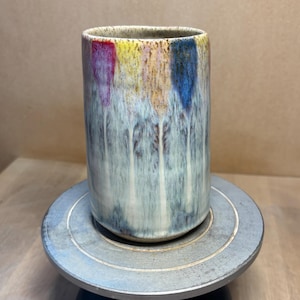 Può includere: Un bicchiere in ceramica fatto a mano con una smaltatura unica. La parte superiore del bicchiere presenta strisce verticali rosse, gialle e blu. La smaltatura cola lungo i lati, creando un effetto strutturato e fluido. Il bicchiere è cilindrico.