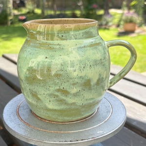 May include: A ceramic jug with a rounded body and a single handle, featuring a speckled light green glaze with darker green and brown accents. The vessel has a wide opening with a small pouring spout and a smooth, glossy finish throughout.