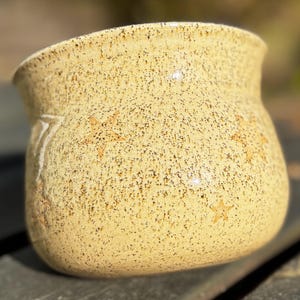 Puede incluir: Una maceta de cerámica amarilla clara moteada, con base redondeada y borde ligeramente ensanchado. La maceta presenta diseños en forma de estrella y una superficie texturizada. Probablemente hecha a mano.