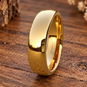May include: A polished gold-tone ring with a smooth, domed exterior surface. The interior of the band features a clear engraving that reads Tungsten Carbide. The ring has a reflective finish that captures light across its curved, solid metal band.