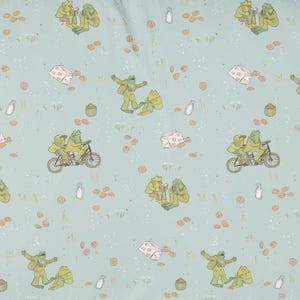 May include: A light blue fabric featuring a repeating pattern of green frogs in various activities, including riding a tandem bicycle, reading books, and dancing, interspersed with small illustrations of milk cartons and scattered brown cookies.