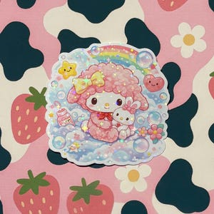 May include: A holographic sticker featuring a pink cartoon character with a yellow bow, a bunny, and a rainbow. The sticker is surrounded by bubbles, stars, and flowers. The background is a pink cow print with strawberries and daisies.