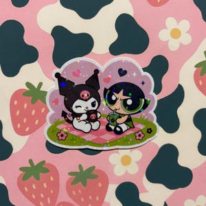 May include: A holographic sticker featuring the character Kuromi and Buttercup from The Powerpuff Girls sitting on a pink and white checkered picnic blanket. The design includes small hearts, flowers, and green grass with a sparkling star effect.