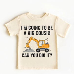 Puede incluir: Camiseta crema para niños pequeños con el texto "I'M GOING TO BE A BIG COUSIN CAN YOU DIG IT?" y una excavadora de dibujos animados en naranja y amarillo. La excavadora está recogiendo un montón de tierra gris.